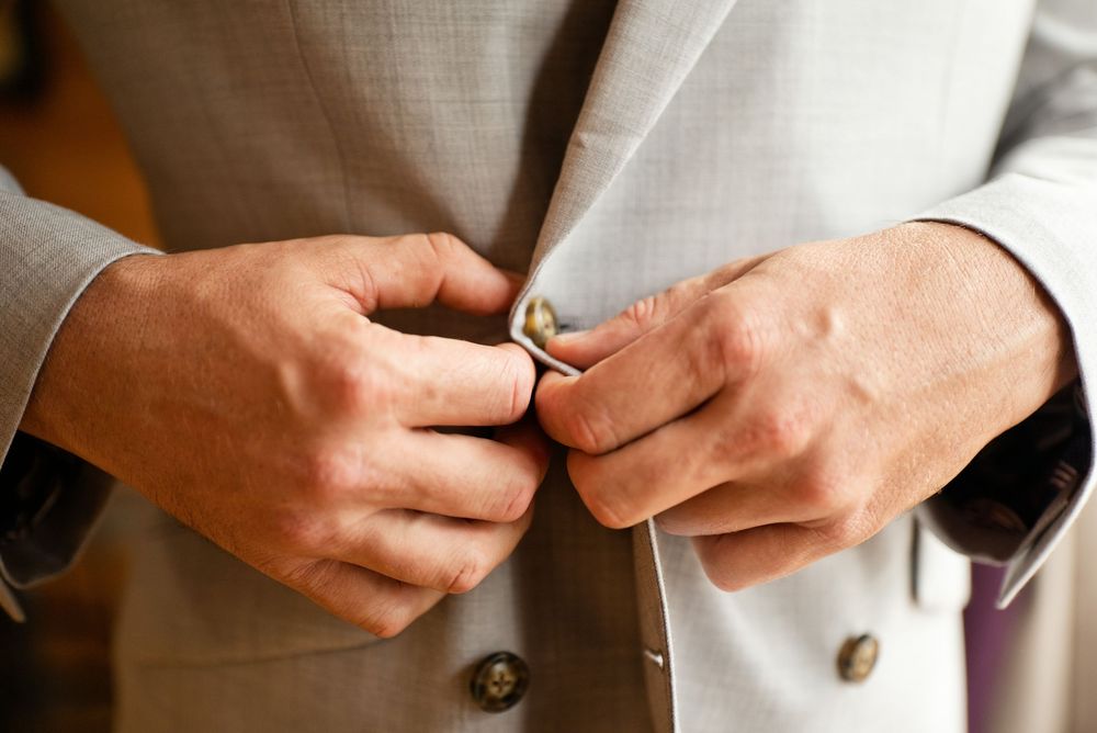 Man in light gray suit jacket buttoning his coat, wearing a ring on his finger.