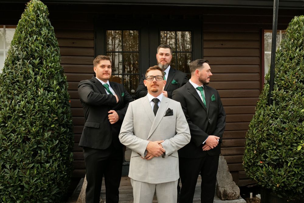 Groom in light gray suit stands with three groomsmen in black suits and teal bow ties outside a wooden venue.