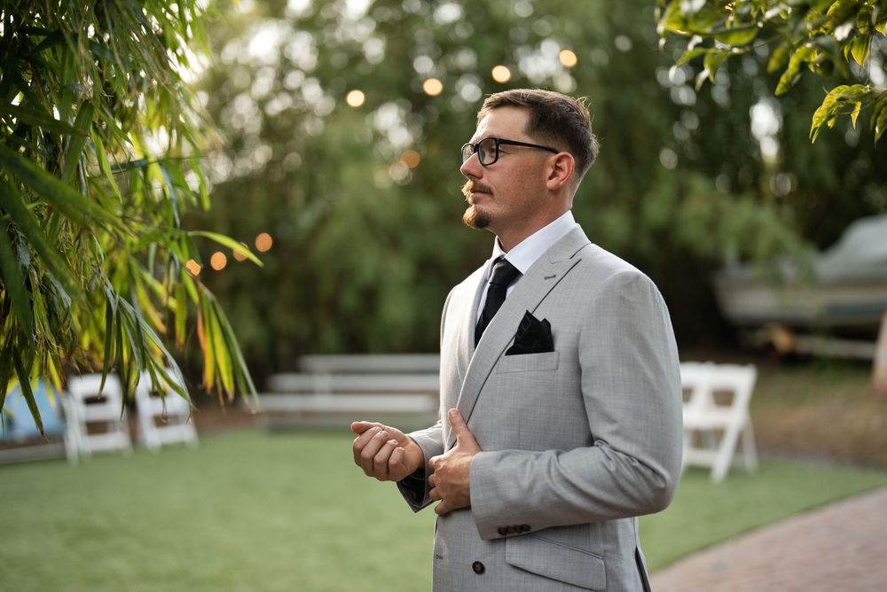 A well-dressed man in a light gray suit and black tie stands outdoors at a garden wedding venue.