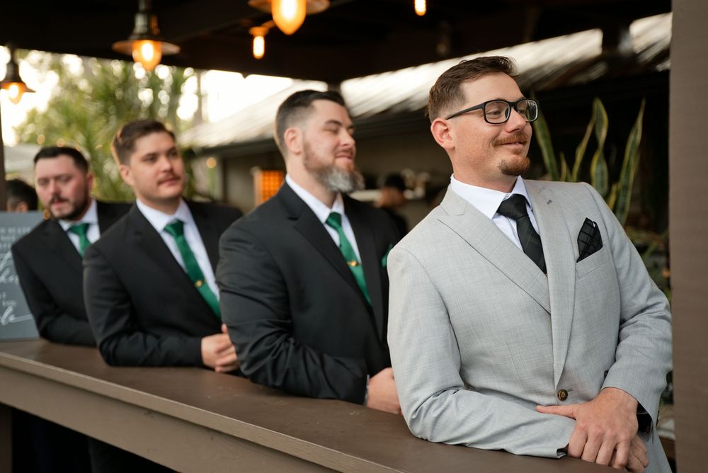 Groom in light gray suit with groomsmen in dark suits and green ties at an outdoor wedding ceremony.