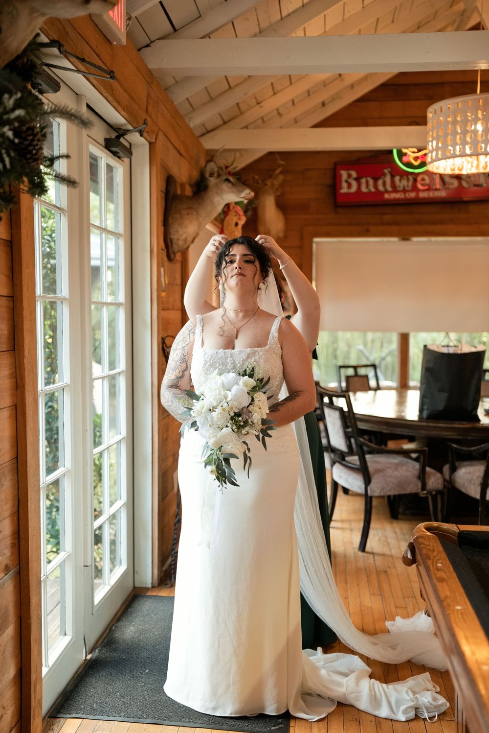 Bride in white gown holds veil overhead, bouquet in hand, standing by French doors in rustic wood-paneled venue.