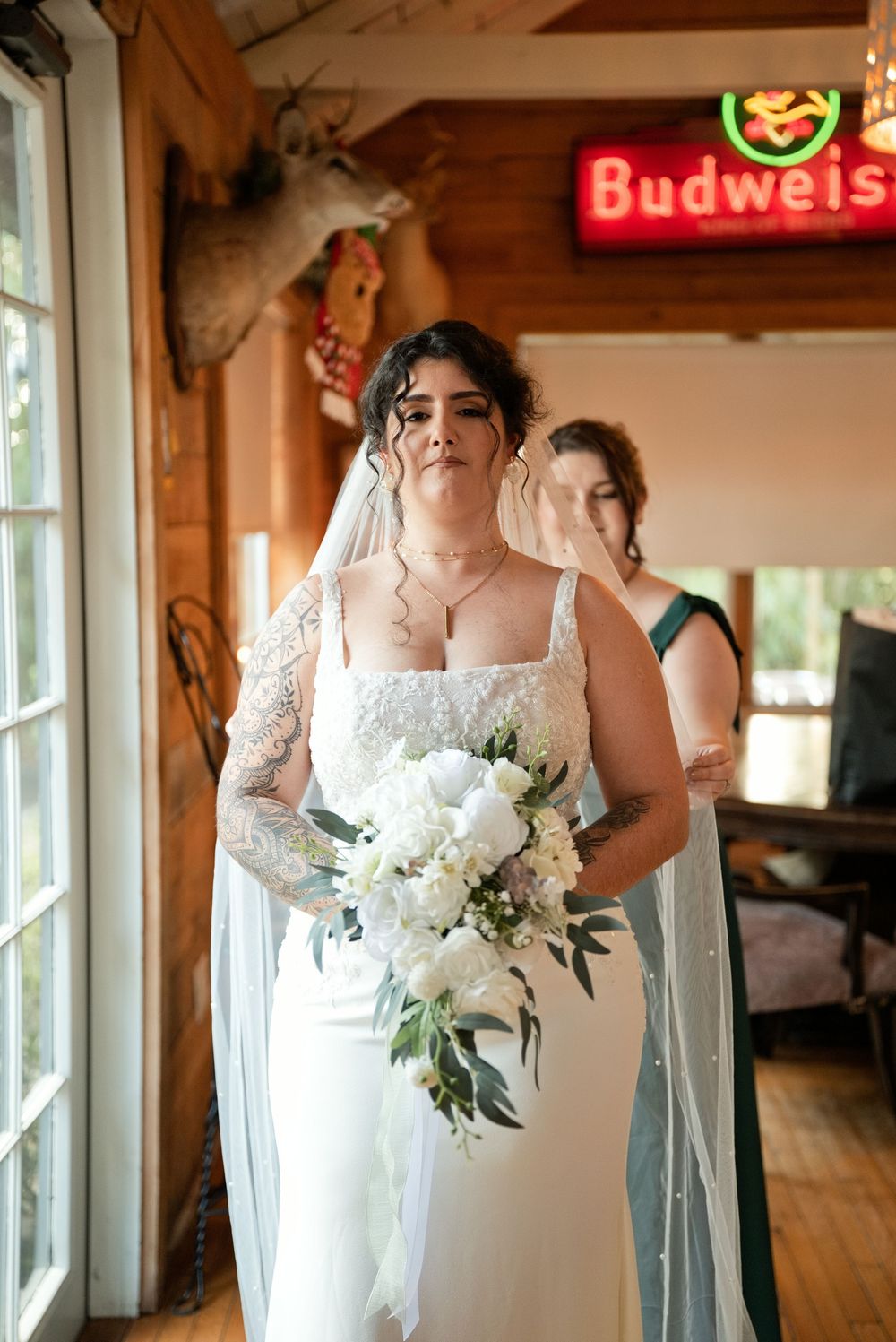 Bride in white gown holding white floral bouquet with veil, standing indoors near Budweiser sign with bridesmaid behind her.