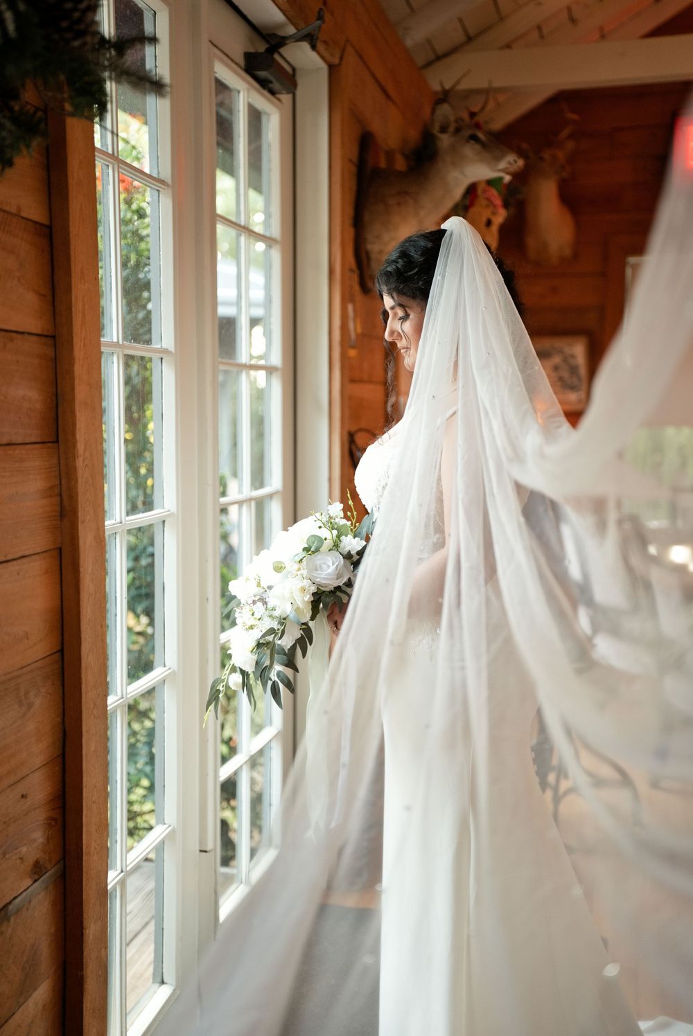 Bride in white gown and flowing veil holds bouquet by sunlit window in rustic wood-paneled room.