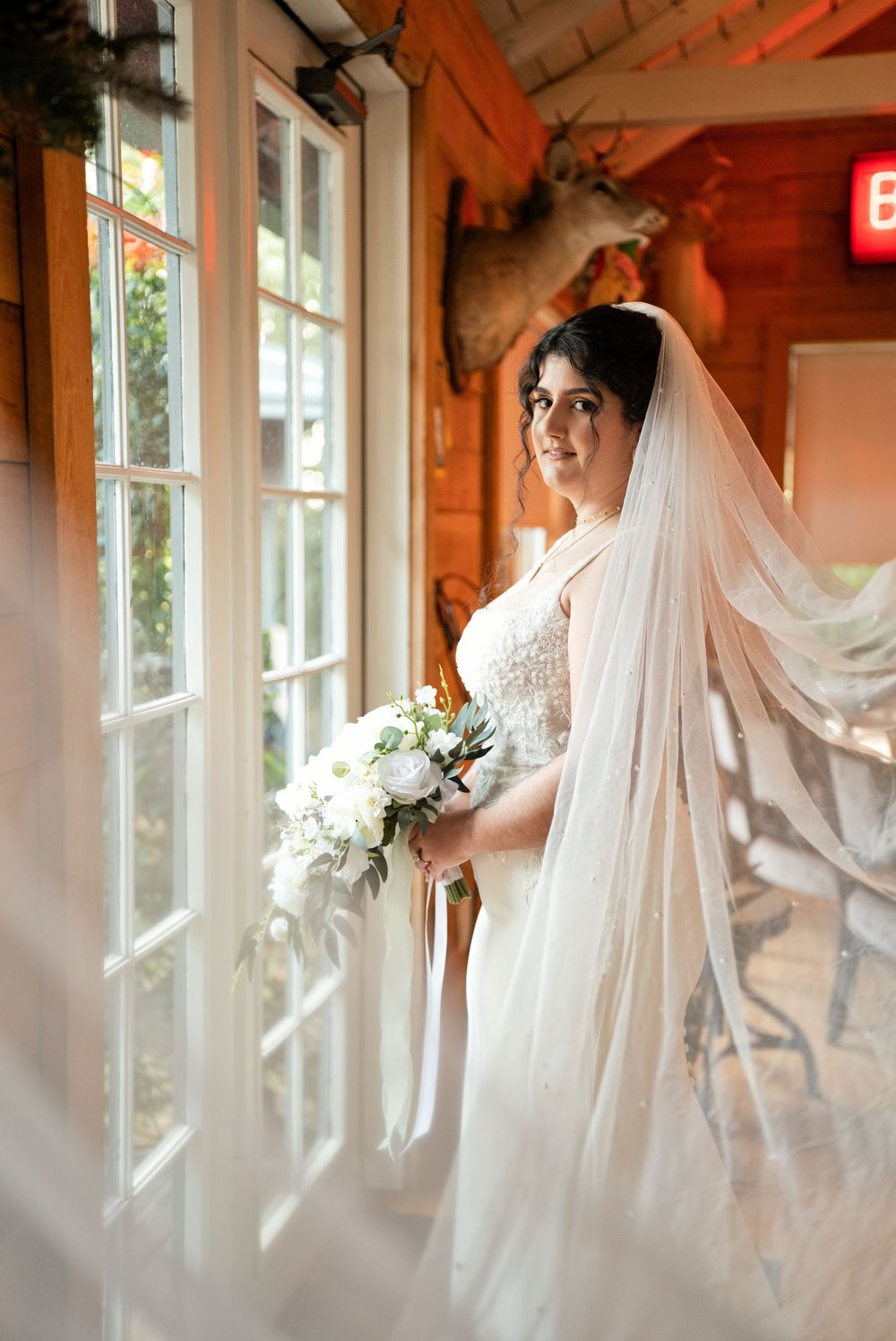 Bride in white lace gown and flowing veil holds white bouquet by sunlit windows in rustic venue.