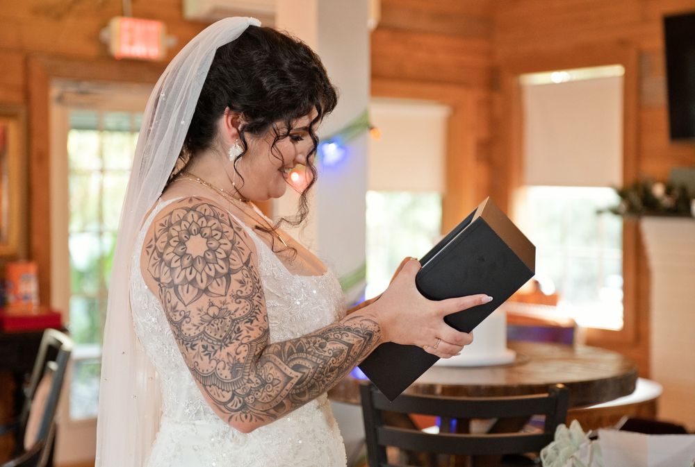 Tattooed bride in white wedding dress and veil reading vows from a black book indoors.