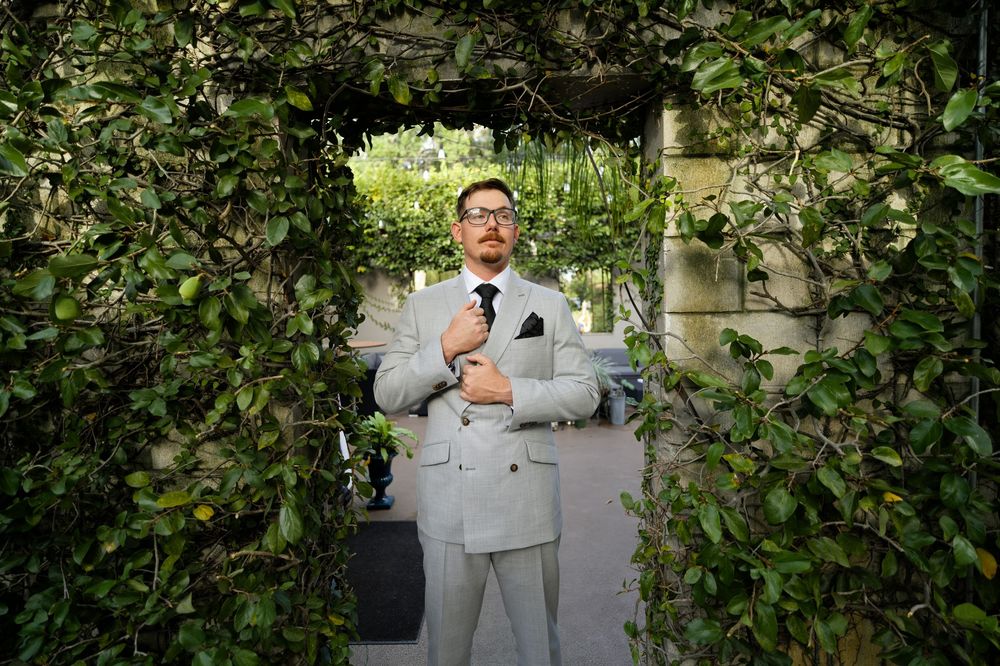 Man in light gray double-breasted suit adjusting tie under lush green vine archway outdoors.