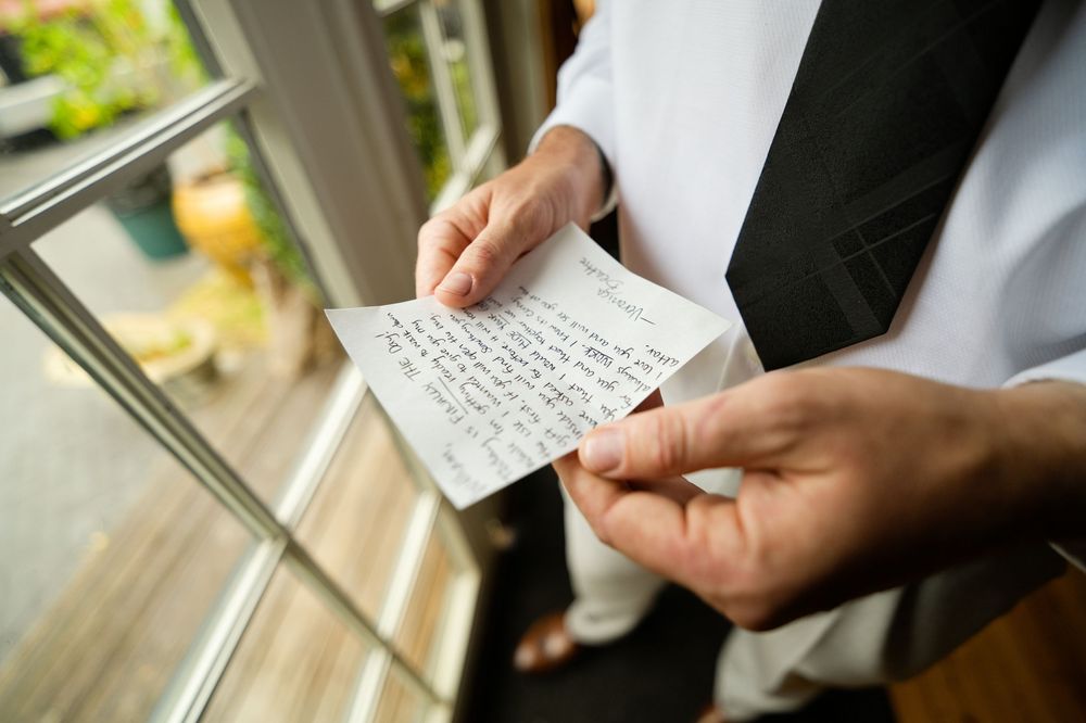 Groom in white shirt and black vest reading a handwritten note card by a window on wedding day.