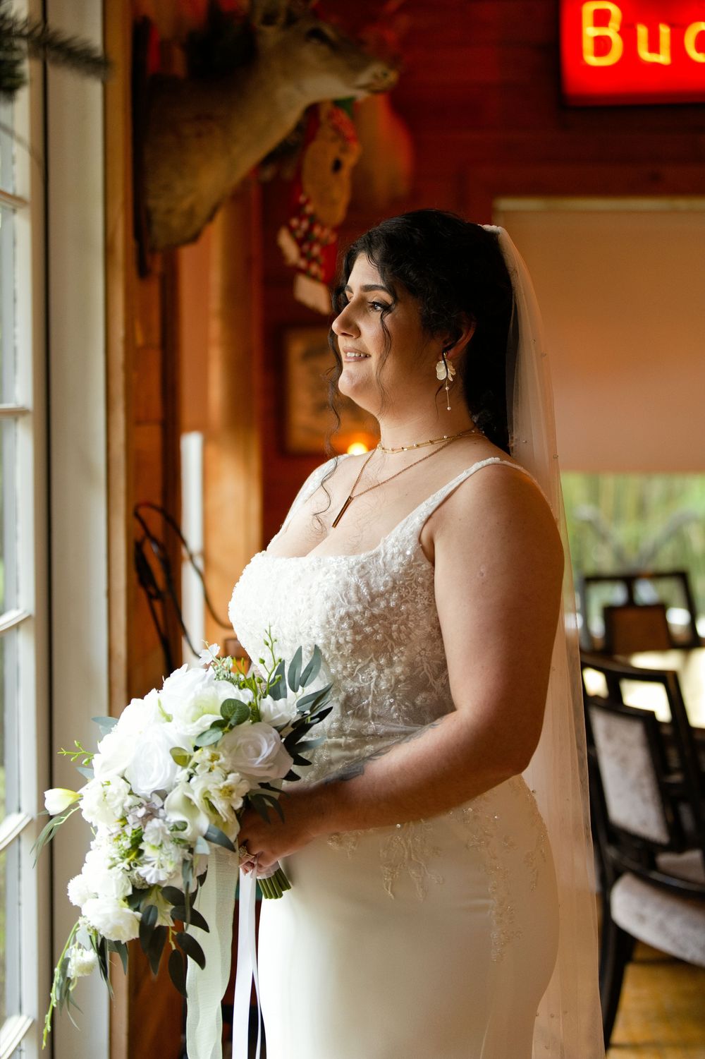 Bride in white lace gown holding bouquet of white flowers, standing by window wearing veil and jewelry.