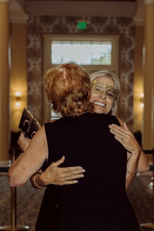 Two women in black dresses share a warm embrace at a masquerade ball event.