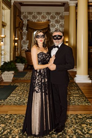 Elegant couple in formal attire wearing black masquerade masks posing in ornate ballroom hallway.