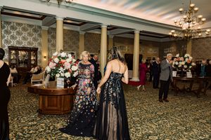 Two women in elegant black floral gowns converse at a formal gala event in an ornate ballroom.