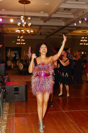 Energetic female performer in colorful feathered dress singing on a ballroom dance floor at a formal gala event.