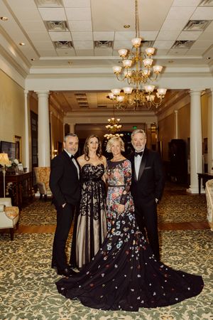 Two couples in elegant formal attire pose in a grand ballroom with chandeliers and ornate decor.