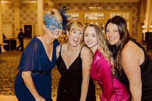 Four women posing together at a formal gala, one wearing a decorative masquerade mask in an elegant ballroom setting.