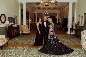 Three elegantly dressed guests posing in grand hotel hallway with ornate decor and patterned carpet.
