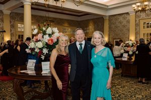 Three elegantly dressed guests pose together at a formal gala event in a decorated ballroom.