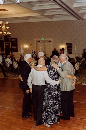 Group of seniors share a warm embrace while dancing together on a ballroom dance floor.