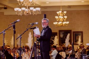 Formally dressed emcee in tuxedo addresses a large crowd at an elegant ballroom gala.