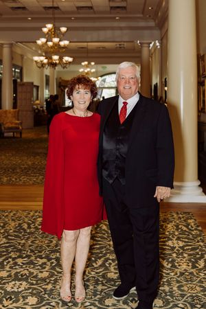 Elegant couple posing in formal attire, woman in red dress and man in black suit with red tie, in a grand hotel ballroom.
