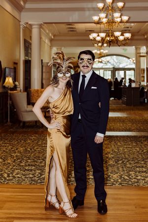 Elegant couple in gold dress and black suit wearing masquerade masks at a formal gala.