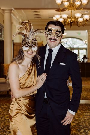 Couple in elegant masquerade masks and formal attire posing at a glamorous ballroom event.