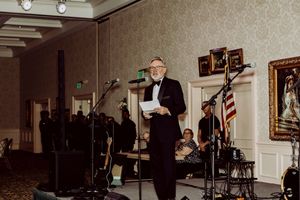 Man in tuxedo speaks at podium during formal event with band and American flag in elegant ballroom.