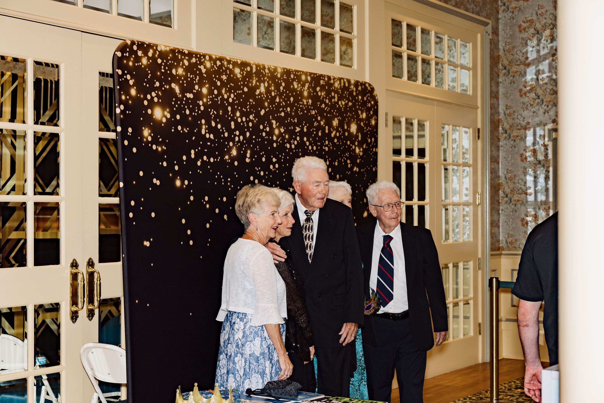 Elderly guests socializing in front of a sparkling black backdrop at an elegant indoor event.