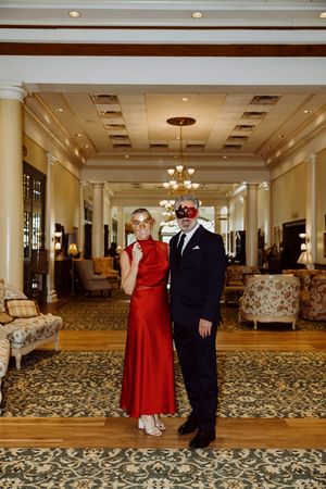 Couple in formal attire wearing decorative masquerade masks pose in a grand hotel ballroom lobby.