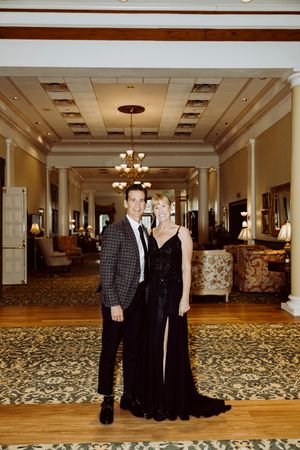 Elegant couple poses in hotel lobby, man in plaid blazer and woman in black velvet gown.