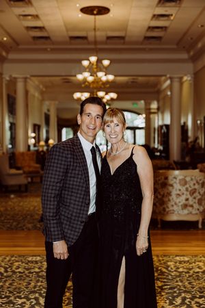 Smiling couple in formal black attire poses in a grand hotel lobby with chandelier backdrop.