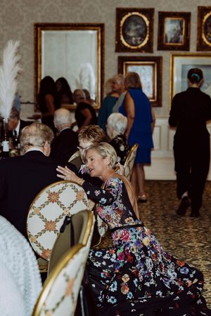 Guests in formal attire socializing at an elegant event, woman in floral gown conversing at a decorated table.