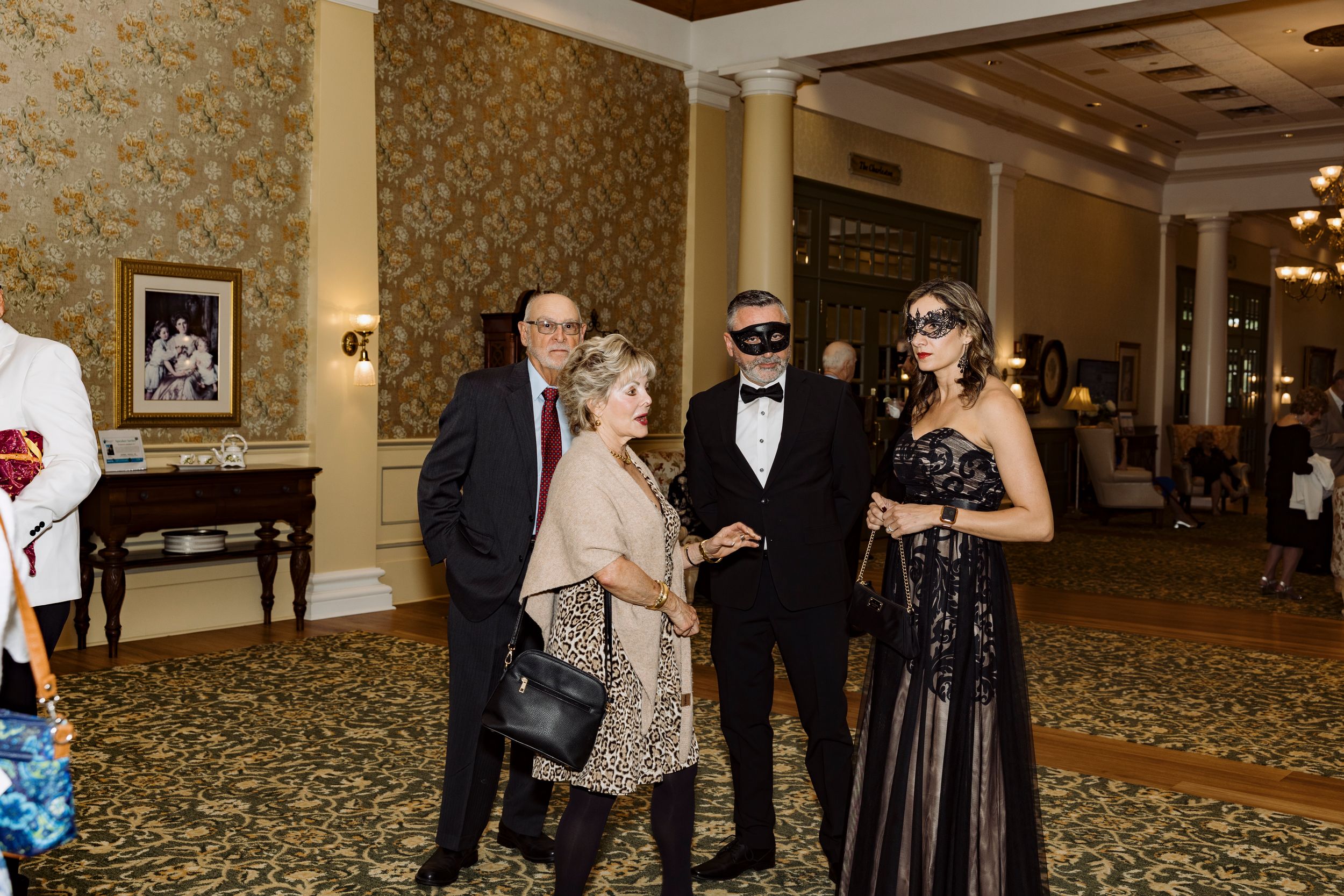 Guests in elegant attire and masquerade masks mingle at a formal gala event in a grand ballroom.