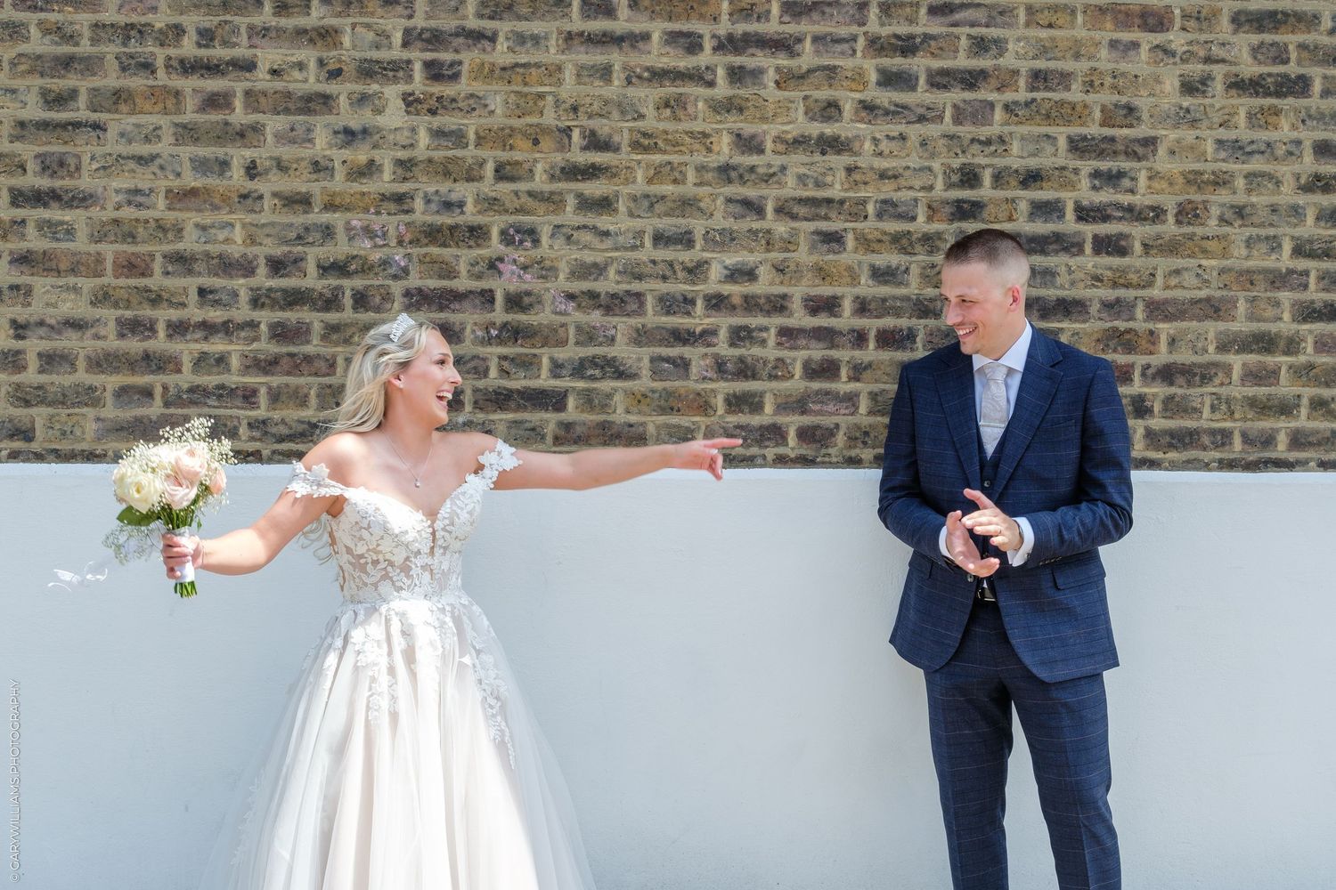 Bride in white gown laughing and reaching toward groom in navy suit outdoors against brick wall on wedding day.