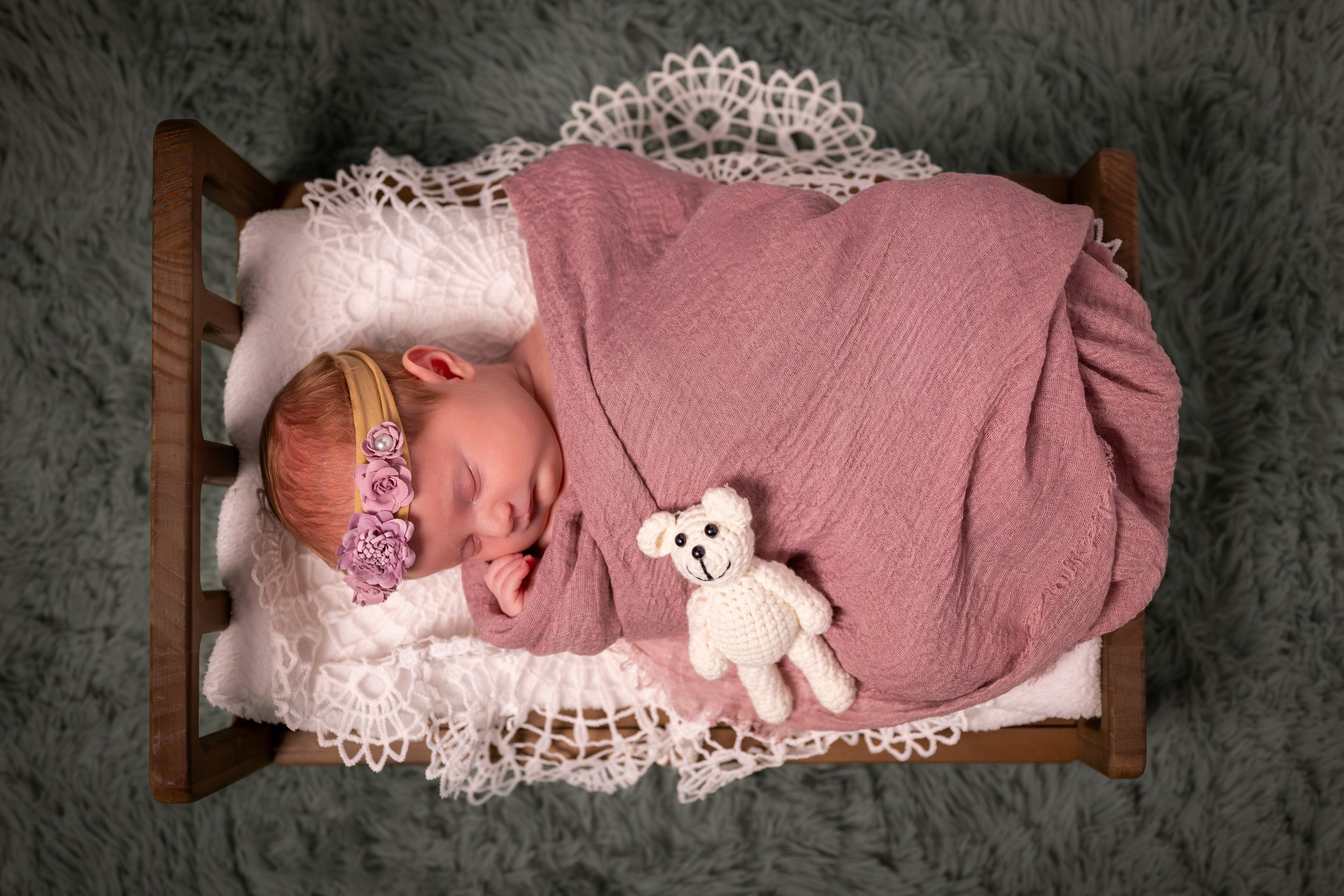 Sleeping newborn baby wrapped in pink swaddle with teddy bear in small wooden bed with lace blanket.