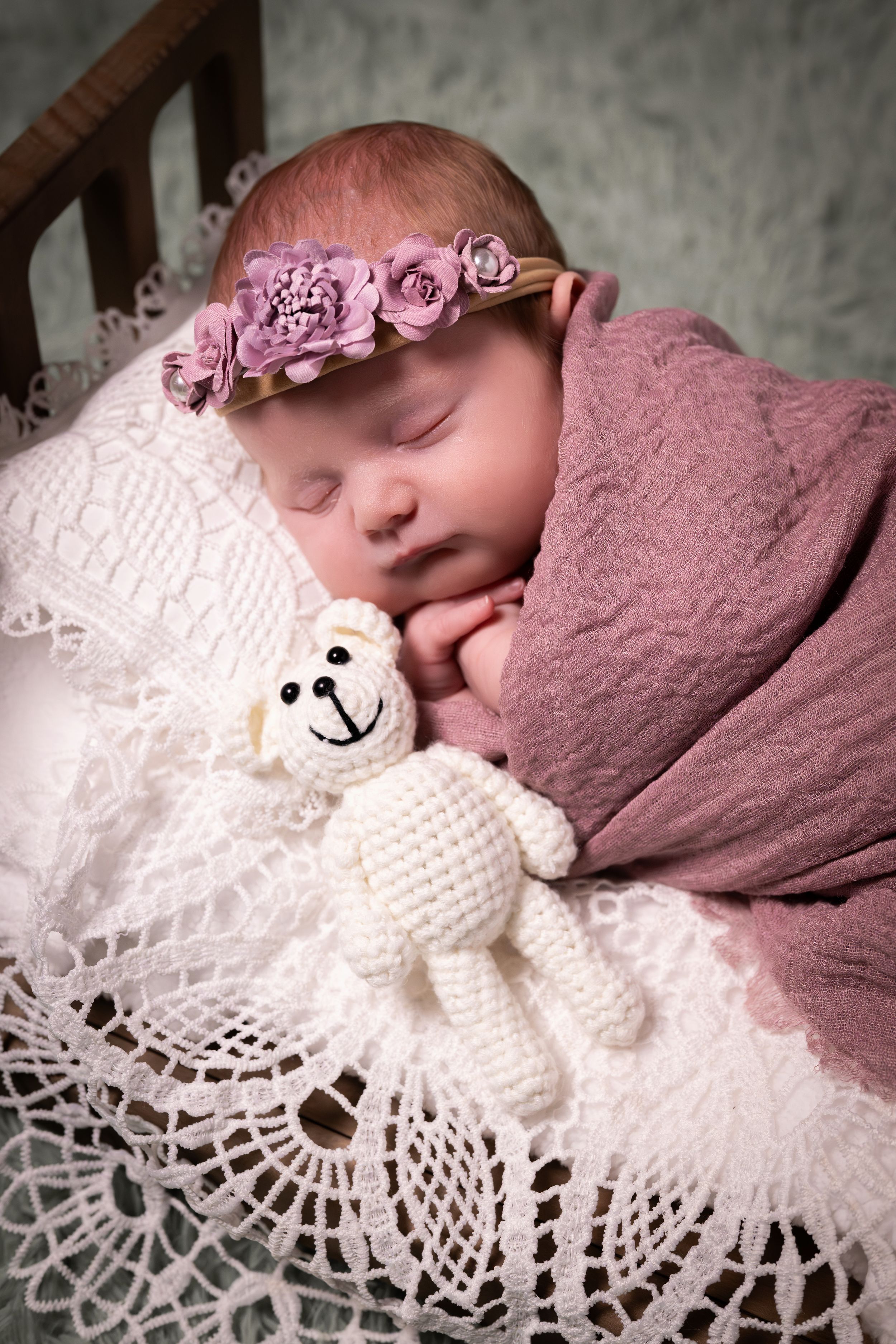 Sleeping newborn baby wrapped in pink with floral headband cuddling a crochet teddy bear on lace blanket.