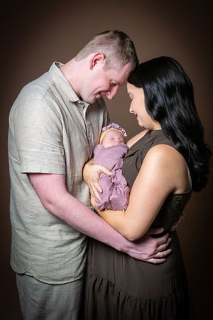 New parents touch foreheads while holding their purple-swaddled newborn in a warm studio photo.
