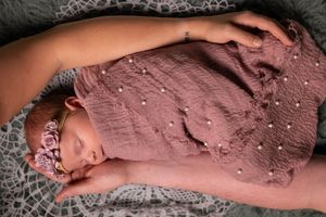 Newborn baby sleeping peacefully wrapped in a pink muslin swaddle with pearl details and floral headband.