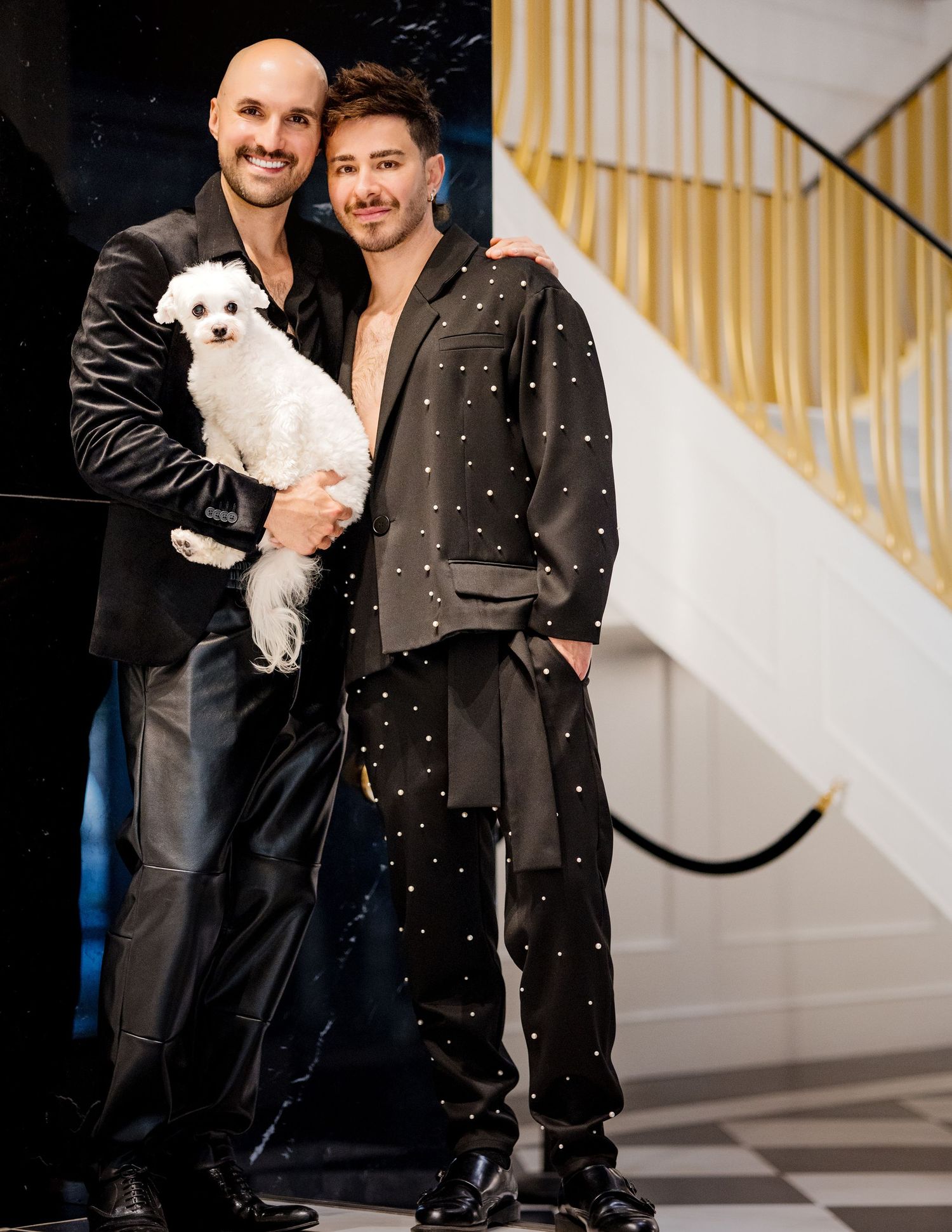 Two men in elegant black outfits pose on staircase, one holding a small white fluffy dog.