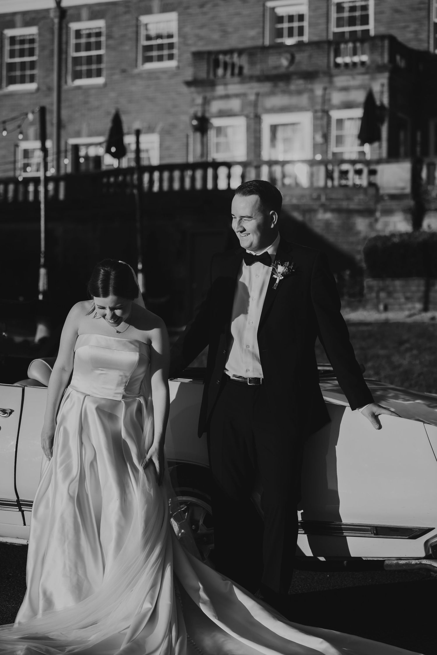 Black and white photo of a bride and groom standing by a vintage car outside an elegant brick building.