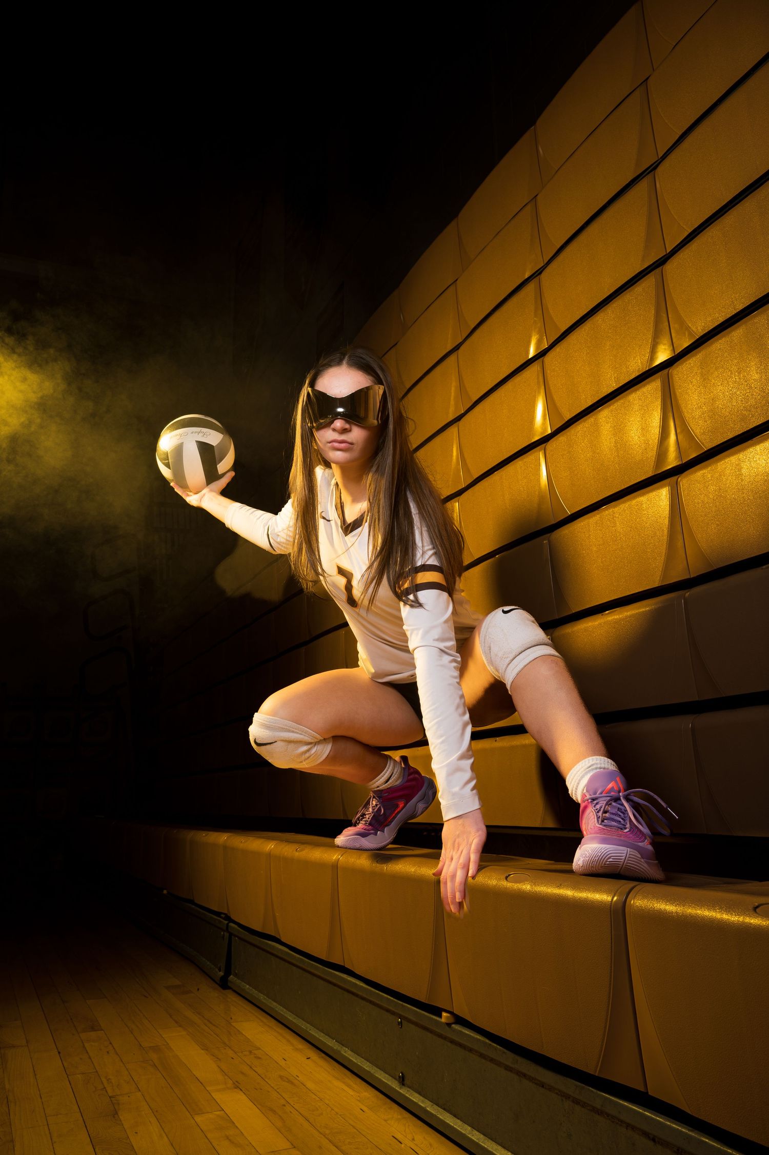 Female volleyball player in white outfit and sunglasses crouching on golden bleachers holding a volleyball.