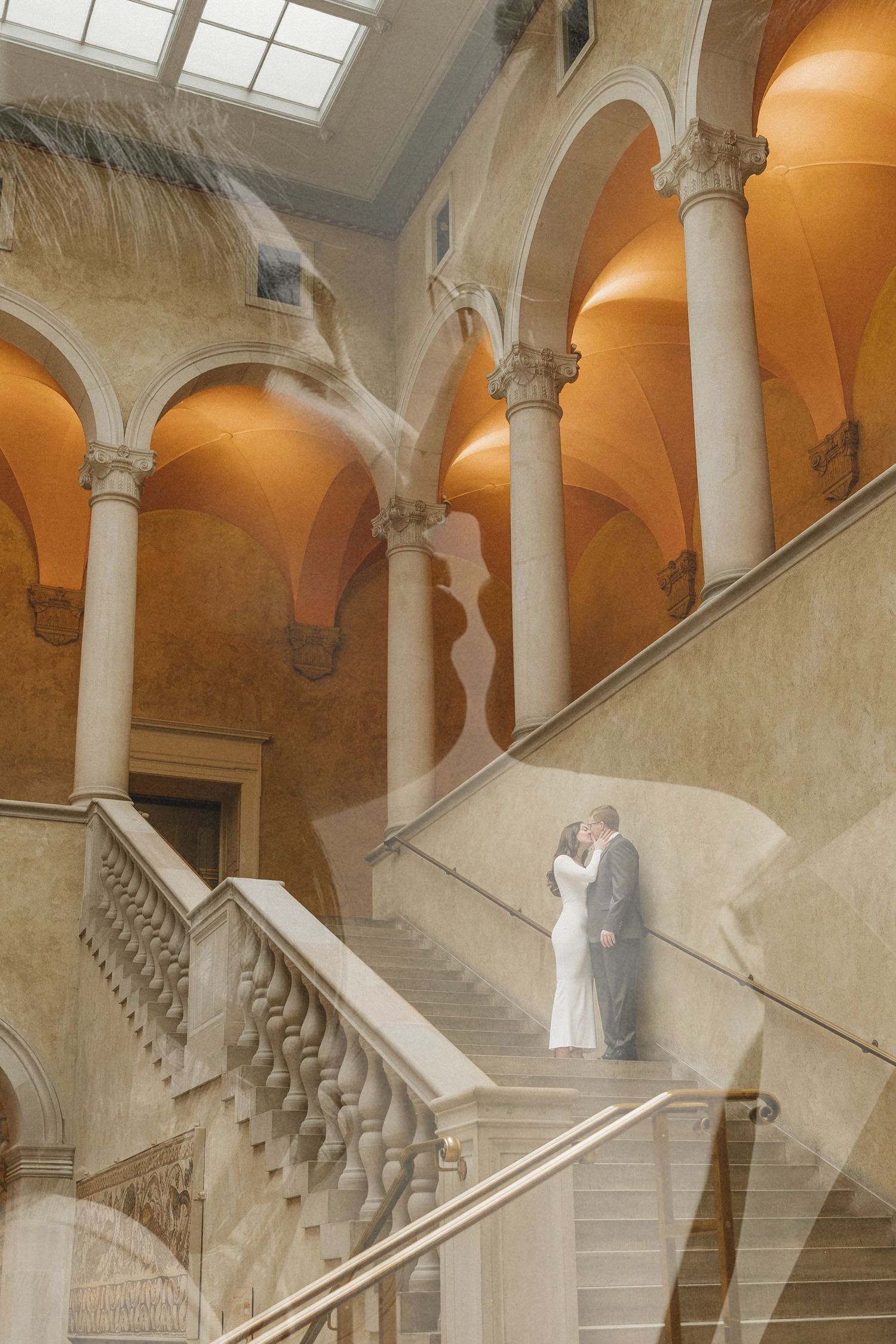 Bride and groom share a romantic moment on grand marble staircase in elegant arched hall with ornate columns.
