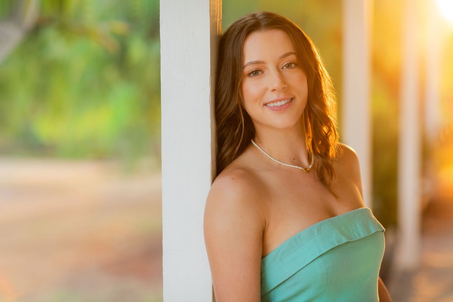 Young woman in teal strapless dress smiling warmly, leaning against white column with golden sunlit greenery behind her.