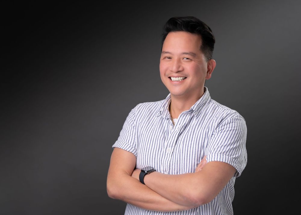 Smiling Asian man in striped shirt with arms crossed poses confidently against dark gray background.