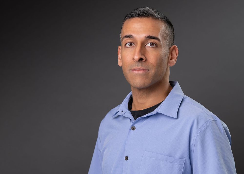 Professional headshot of a man in a light blue button-up shirt against a gray background.