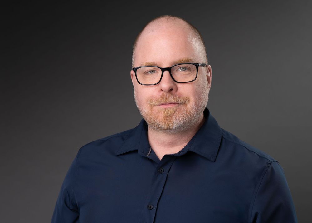 Professional headshot of a bald man with glasses and a beard wearing a navy blue shirt against a gray background.