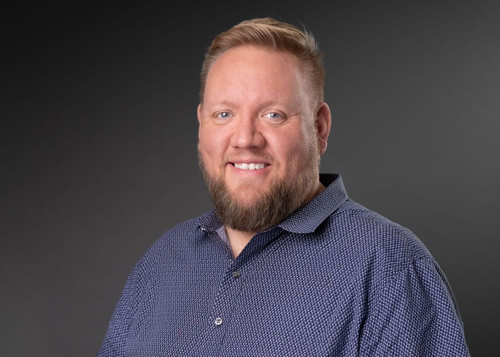Professional headshot of a smiling man with a blonde beard wearing a navy patterned shirt against a gray background.