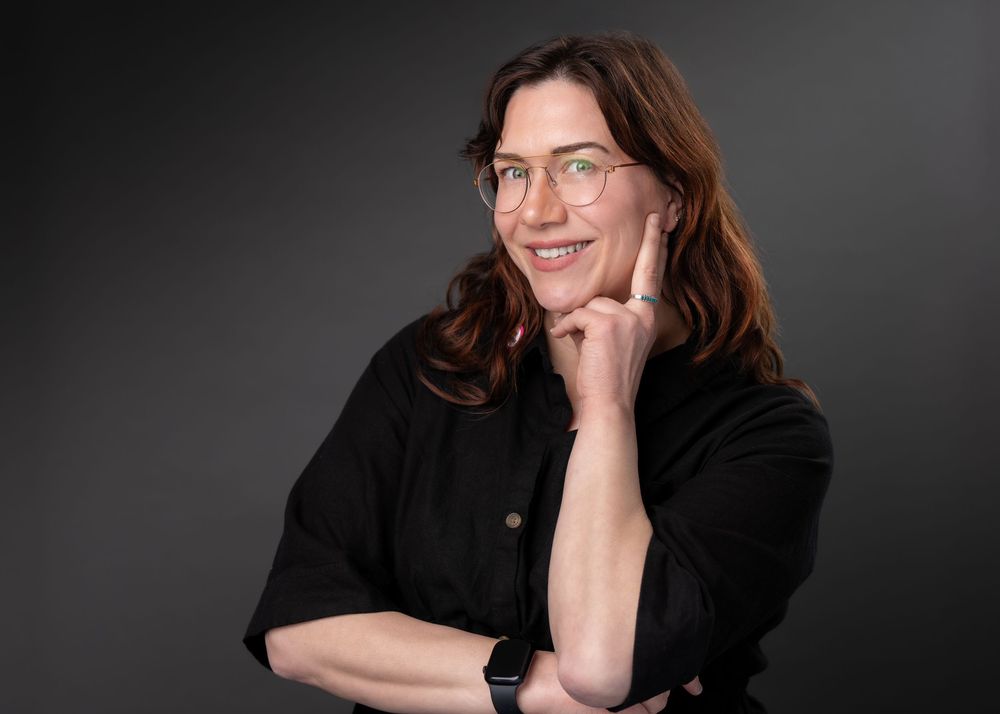 Smiling woman with brown hair and glasses poses thoughtfully with hand on chin against dark background.
