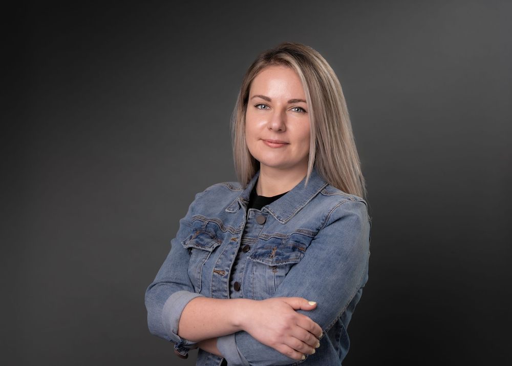 Professional headshot of a blonde woman in a denim jacket with arms crossed against a dark background.