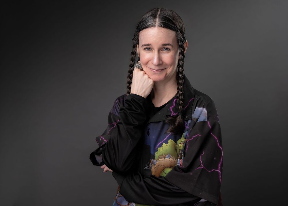 Woman with braided pigtails wearing a black graphic sweatshirt, posing confidently against a gray background.