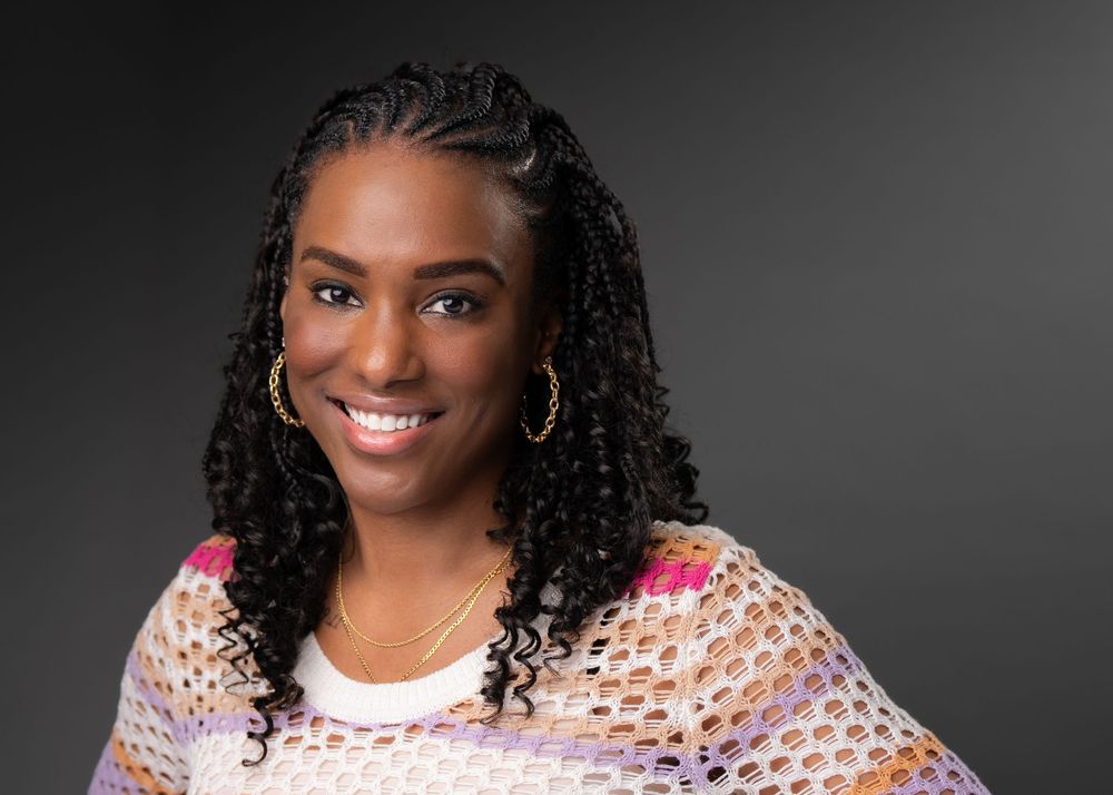 Professional headshot of a smiling woman with braided hair wearing a colorful crochet shawl against a dark background.